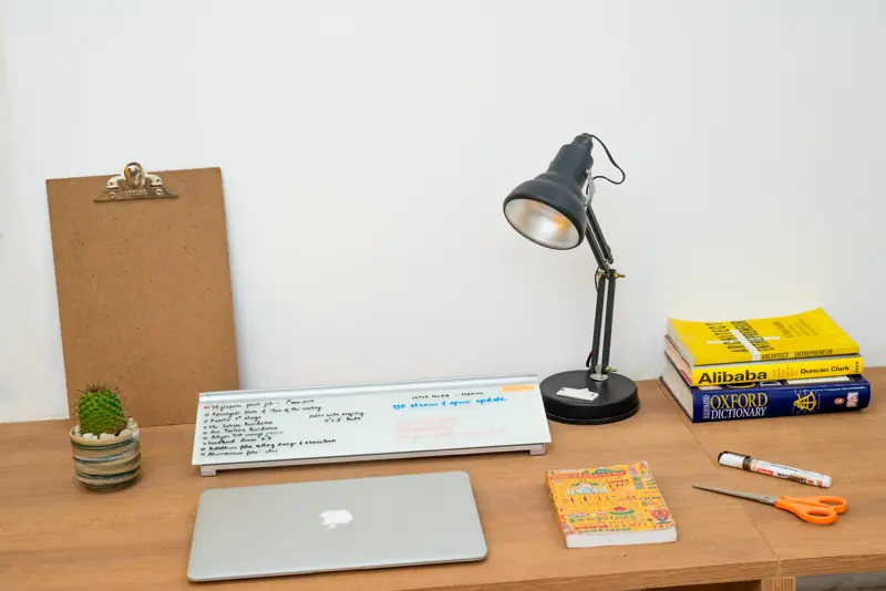 A wooden desk with a laptop, an anglepoise lamp, books and a small cactus
